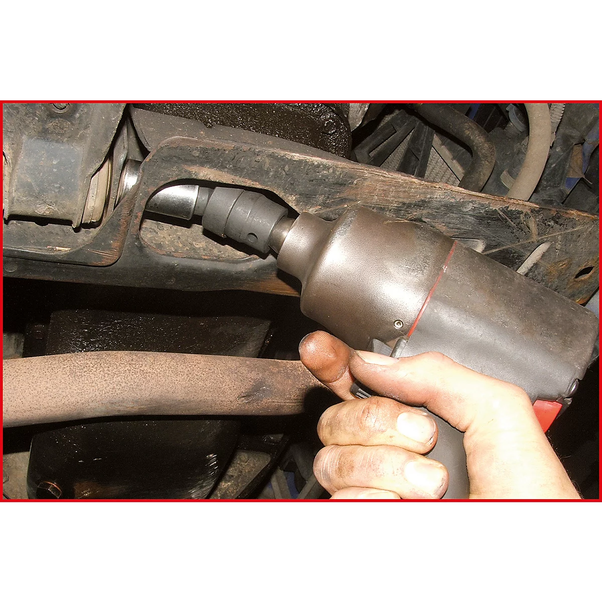 Eine Hand mit öligen Fingern hält einen Schlagschrauber, der auf ein Autoteil gerichtet ist. Rostige Rohre und Fahrzeugteile im Hintergrund.